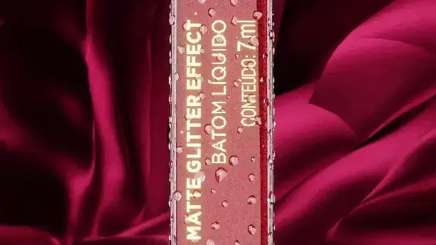 Batom líquido Avon Matte Glitter Effect em still de vídeo publicitário, sobre fundo de cetim vermelho com gotas d'água